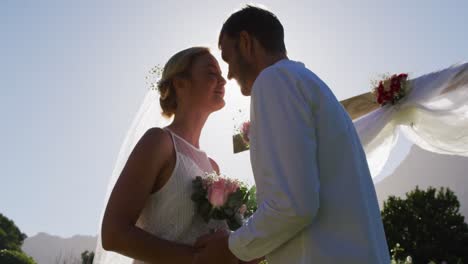 Happy-caucasian-newly-wed-couple,-standing-touching-heads-at-altar-outdoors