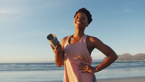 Portrait-of-african-american-woman-holding-yoga-mat-at-the-beach-smiling