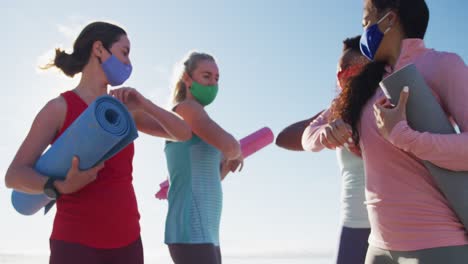 Gruppe-Verschiedener-Freundinnen-Mit-Gesichtsmasken,-Die-Yogamatten-Am-Strand-Halten-Und-Ihre-Ellbogen-Berühren