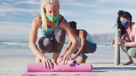 Gruppe-Verschiedener-Freundinnen-Mit-Gesichtsmasken,-Die-Am-Strand-Yogamatten-Rollen