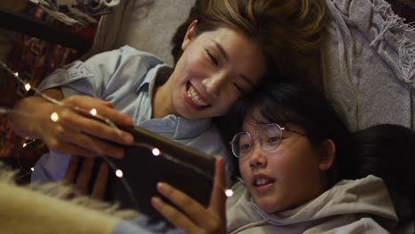 Asiatische-Mutter-Und-Tochter-Lächeln,-Während-Sie-Zu-Hause-Unter-Einer-Deckenfestung-Liegen-Und-Ein-Digitales-Tablet-Benutzen
