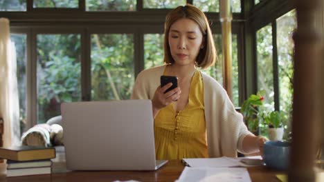Asiatische-Frau-Sitzt-Am-Tisch,-Arbeitet-Von-Zu-Hause-Aus-Und-Nutzt-Das-Smartphone