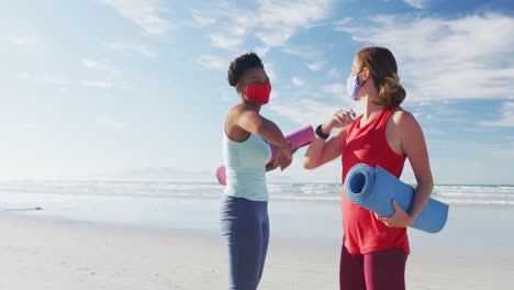 Two-diverse-female-friends-wearing-face-masks-holding-yoga-mats-at-the-beach-touching-elbows