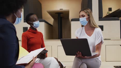 Three-two-diverse-female-office-colleagues-wearing-face-masks-discussing-together-at-office