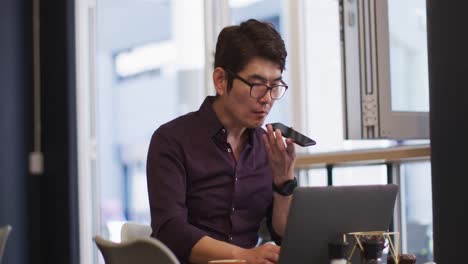 Asiatischer-Mann-Mit-Laptop,-Der-Auf-Dem-Smartphone-Spricht,-Während-Er-In-Einem-Café-Sitzt
