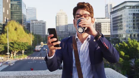 Hombre-Asiático-Tomando-Café-Y-Usando-Un-Teléfono-Inteligente-Mientras-Está-Sentado-En-El-Techo-Del-Parque-Corporativo