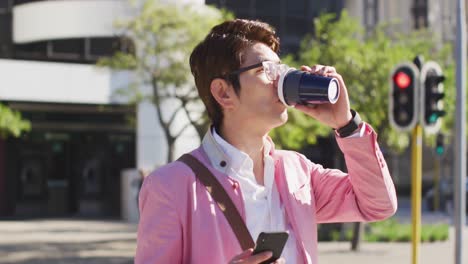 Asiatischer-Mann-Trinkt-Kaffee-Und-Benutzt-Smartphone,-Während-Er-Die-Straße-überquert