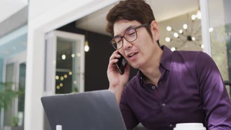 Asiatischer-Mann-Spricht-Auf-Smartphone-Und-Benutzt-Laptop,-Während-Er-In-Einem-Café-Sitzt