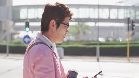 Hombre-Asiático-Tomando-Café-Y-Usando-Un-Teléfono-Inteligente-Mientras-Camina-Por-La-Calle