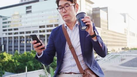 Asiatischer-Mann-Trinkt-Kaffee-Und-Benutzt-Smartphone,-Während-Er-Auf-Dem-Dach-Des-Unternehmensparks-Sitzt