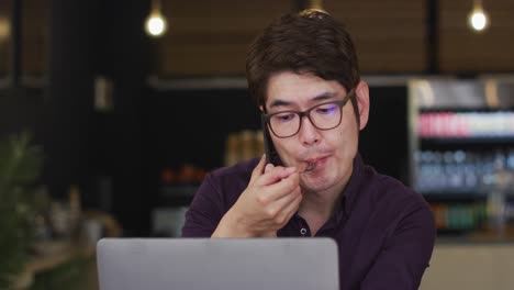 Hombre-Asiático-Con-Una-Computadora-Portátil-Tomando-Un-Refrigerio-Y-Hablando-Por-Teléfono-Inteligente-Mientras-Está-Sentado-En-Un-Café
