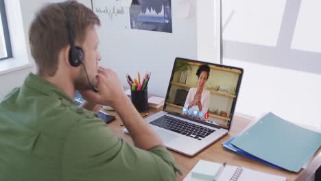 Caucasian-man-wearing-phone-headset-having-a-video-call-with-female-colleague-on-laptop-at-office