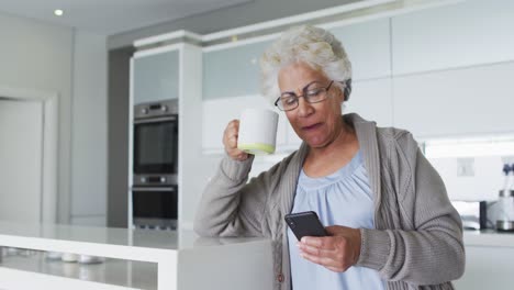 Mujer-Mayor-Afroamericana-Tomando-Café-Y-Usando-Un-Teléfono-Inteligente-En-La-Cocina-De-Casa