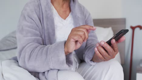 Mittelteil-Einer-Afroamerikanischen-Seniorin,-Die-Ihr-Smartphone-Benutzt,-Während-Sie-Zu-Hause-Auf-Dem-Bett-Sitzt