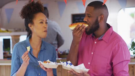 Happy-diverse-couple-talking-and-eating-takeaway-food-by-food-truck