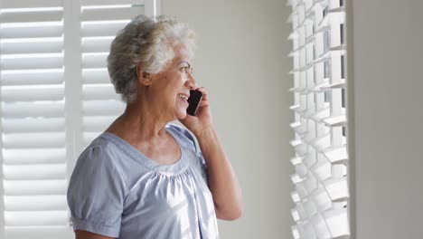 Afroamerikanische-ältere-Frau,-Die-Auf-Dem-Smartphone-Spricht-Und-Zu-Hause-Aus-Dem-Fenster-Schaut