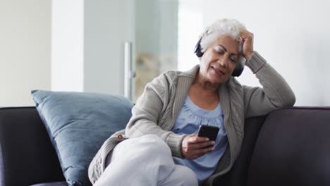 Afroamerikanische-ältere-Frau-Mit-Kopfhörern-Und-Smartphone,-Die-Zu-Hause-Auf-Der-Couch-Sitzt