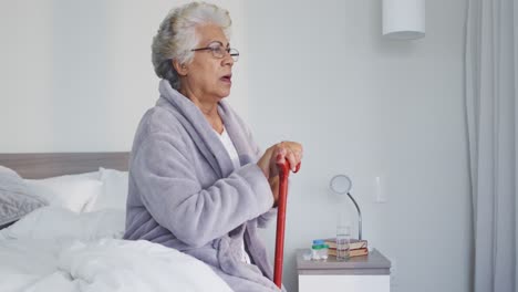 Thoughtful-african-american-senior-woman-holding-walking-stick-sitting-on-the-bed-at-home