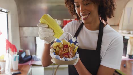 African-american-woman-wearing-apron-putting-mustard-over-the-hot-dog-in-food-truck