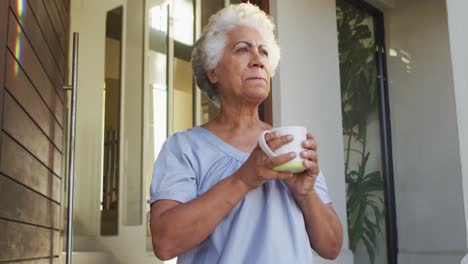 Eine-Afroamerikanische-Seniorin-Trinkt-Kaffee,-Während-Sie-Zu-Hause-An-Der-Haustür-Steht
