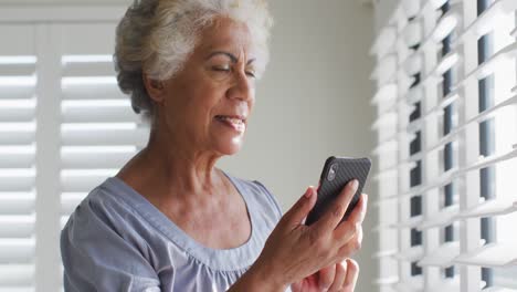 African-american-senior-woman-using-smartphone-and-looking-out-of-the-window-at-home