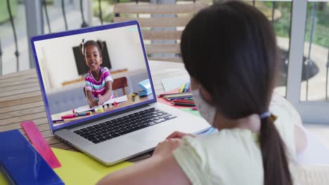 Rückansicht-Eines-Mädchens-Mit-Gesichtsmaske,-Das-Zu-Hause-Einen-Videoanruf-Mit-Einem-Afroamerikanischen-Mädchen-Auf-Einem-Laptop-Führt