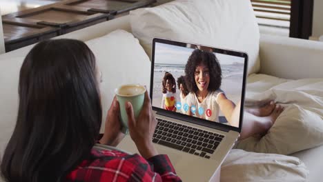 Eine-Afroamerikanische-Frau-Hält-Eine-Kaffeetasse-In-Der-Hand-Und-Führt-Einen-Videoanruf-Auf-Einem-Laptop,-Der-Zu-Hause-Auf-Der-Couch-Sitzt
