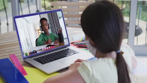 Rückansicht-Eines-Mädchens-Mit-Gesichtsmaske,-Das-Zu-Hause-Einen-Videoanruf-Mit-Einem-Afroamerikanischen-Jungen-Auf-Einem-Laptop-Führt