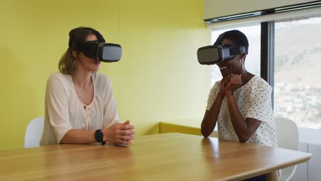 Two-diverse-female-coworkers-sitting-at-desk,-testing-vr-googles