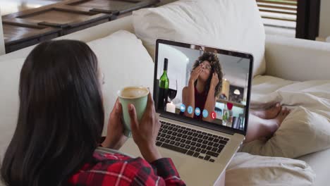 Mixed-race-woman-sitting-on-sofa-using-laptop-making-video-call-with-female-friend