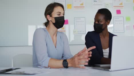 Two-diverse-female-colleagues-wearing-face-mask,-sitting-at-desk,-looking-at-laptop-and-talking