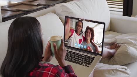 Mixed-race-woman-sitting-on-sofa-using-laptop-making-video-call-with-friends