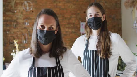 Two-caucasian-women-wearing-face-masks-and-aprons,-looking-at-camera