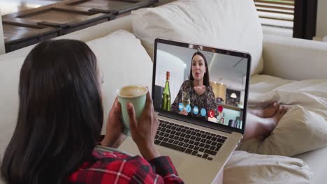 Mixed-race-woman-sitting-on-sofa-using-laptop-making-video-call-with-female-friend