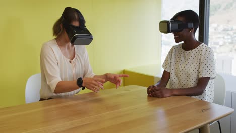 Two-diverse-female-coworkers-sitting-at-desk,-testing-vr-googles
