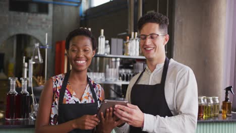 Portrait-of-happy-diverse-couple-wearing-aprons-working-at-a-bar,-using-tablet-and-smiling-to-camera