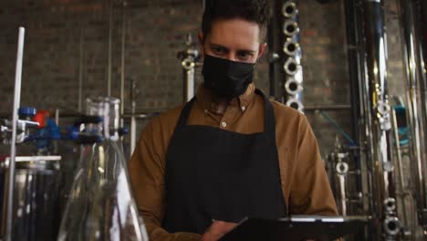 Portrait-of-caucasian-man-working-at-gin-distillery-wearing-face-mask-and-apron-looking-to-camera