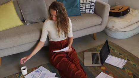Caucasian-woman-working-in-living-room,-sitting-on-floor-with-laptop-and-paperwork,-drinking-coffee