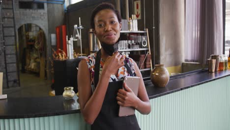 Retrato-De-Una-Mujer-Afroamericana-Con-Mascarilla-Trabajando-En-Un-Bar,-Usando-Una-Tableta-Y-Sonriendo-A-La-Cámara
