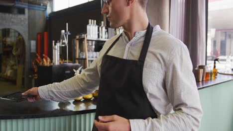 Portrait-of-caucasian-man-wearing-apron-working-at-a-bar,-using-tablet-and-smiling-to-camera