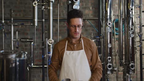 Portrait-of-happy-caucasian-man-working-at-gin-distillery-using-equipment-and-smiling-to-camera