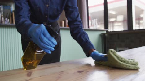 Midsection-of-person-wearing-gloves-and-apron-disinfecting-tables-at-cafe-bar