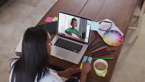 Mixed-race-female-teacher-sitting-at-table-using-laptop-having-online-school-lesson