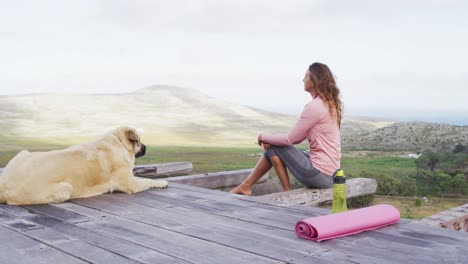 Caucasian-woman-sitting-outdoors-on-deck-with-pet-dog-admiring-view-in-rural-mountainside-setting