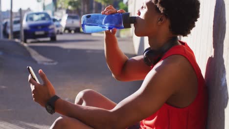 Fit-african-american-man-exercising-in-city-taking-a-break,-using-smartphone,-sitting-drinking-water