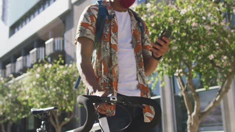 Midsection-of-african-american-man-in-city,-wheeling-bike-and-using-smartphone-in-the-street