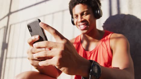 Feliz-Hombre-Afroamericano-En-La-Ciudad,-Usando-Un-Teléfono-Inteligente-Con-Auriculares-Inalámbricos,-Sentado-Contra-La-Pared