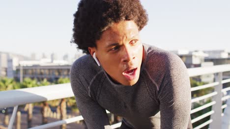 Fit-african-american-man-exercising-outdoors-in-city,-with-wireless-earphones,-resting-on-footbridge