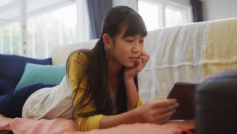 Asiatisches-Mädchen-Lächelt-Und-Benutzt-Ein-Tablet,-Das-Zu-Hause-Auf-Dem-Sofa-Liegt