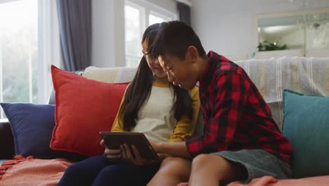 Asian-brother-and-sister-smiling-and-using-tablet-sitting-on-sofa-at-home